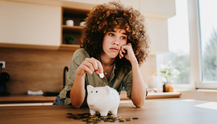 Young woman looking worried while placing coins into a piggy bank Email